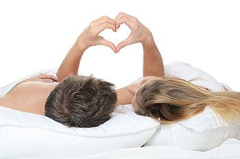 A man and a woman are lying side-by-side in bed, covered by white bedding.
