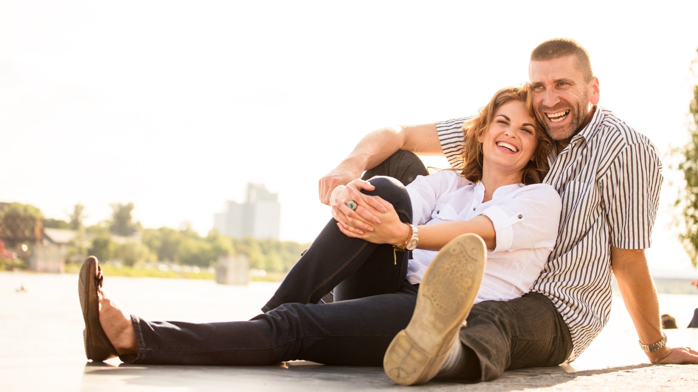 Two individuals are smiling and sitting closely together on the ground. One is wearing a white shirt and blue jeans, while the other is in a striped shirt and dark pants.