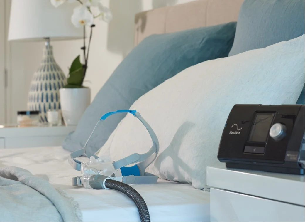 The image shows a close-up of a neatly made bed with white and light blue bedding. On the bed, there is a CPAP mask connected to a hose, which is part of a CPAP machine placed on the bedside table. 